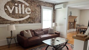 A living room with a brown leather sofa, a glass coffee table, and a patterned rug. A large wall clock and a fireplace are visible in the background.