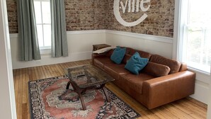 A living room with a brown leather sofa, a glass coffee table, a patterned rug, a brick wall, and a large window with curtains.