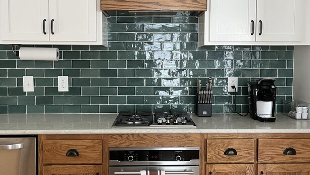 A modern kitchen with white cabinets, a wooden hood, green-tiled backsplash, stainless steel appliances, and wooden lower cabinets.