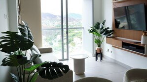 A modern living room with a large TV, a sofa, and a view of a cityscape through a sliding glass door. - Polanco Residences Ambar Roca Santa Marta - Colombia (Santa Marta)