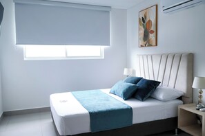 A bedroom with a bed, a nightstand, a lamp, a window with a blind, and a painting on the wall. - Polanco Residences Ambar Roca Santa Marta - Colombia (Santa Marta)