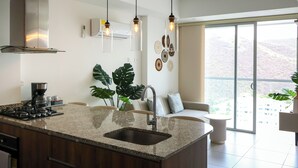 A modern kitchen with a granite countertop, stainless steel appliances, and a large window offering a view of the outdoors. - Polanco Residences Ambar Roca Santa Marta - Colombia (Santa Marta)