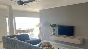 Ein modernes Wohnzimmer mit grauem Sofa, einem Flachbildfernseher, Deckenventilator und Blick ins Freie.