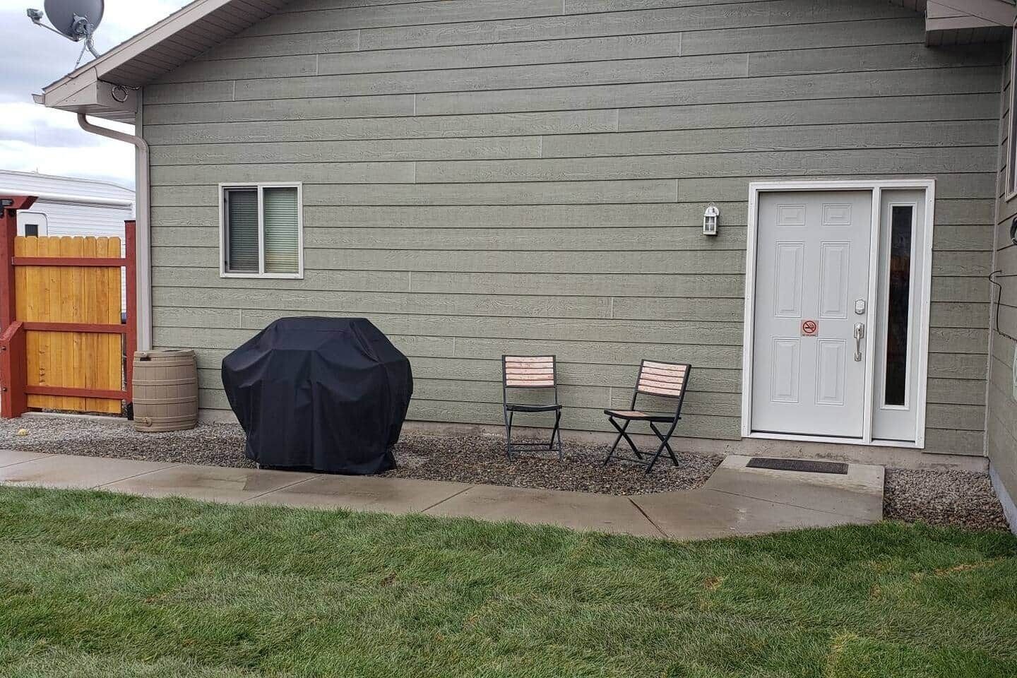 Une maison avec une porte de garage, un barbecue couvert, deux chaises et une porte blanche.
