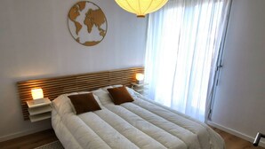 Un lit bien fait, avec une couette blanche et deux oreillers marron, une table de chevet avec une lampe, une carte du monde accrochée au mur et une fenêtre ornée de rideaux légers.