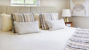A neatly made bed with a beige headboard, several pillows, and a blanket. There is a bedside table with a lamp and a framed picture.