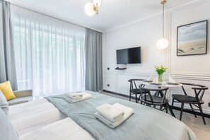 Een hotelkamer met twee bedden, een eettafel met stoelen, een aan de muur gemonteerde televisie en een schilderij van een strandgezicht.