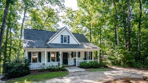 A white house with a gabled roof, surrounded by trees and a gravel driveway. - Orion's Retreat a charming cottage nestled by the scenic shores of Lake Sinclair (Eatonton)