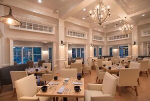 A spacious dining area with multiple tables and chairs, large windows, and elegant chandeliers.