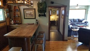 A cozy kitchen with wooden flooring, a bar area with stools, and a dining table. There is a wooden ceiling with hanging lights, a refrigerator, and a shelf with bottles. A framed picture hangs on the wall, and a bear figurine is on the counter. - A Cabin in Rochford (Hill City)