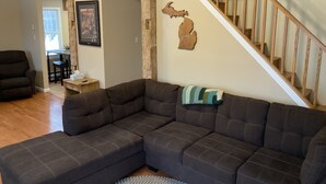A living room with a sectional sofa, a wooden staircase, and a dining area in the background. - Cozy cabin in the woods (Evart)