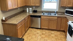 A kitchen with wooden cabinets, a dishwasher, a sink, a microwave, and a window with a view of trees. - Cozy cabin in the woods (Evart)