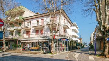 A street view with a building featuring balconies, a visible storefront with outdoor items, and a clear sky.
