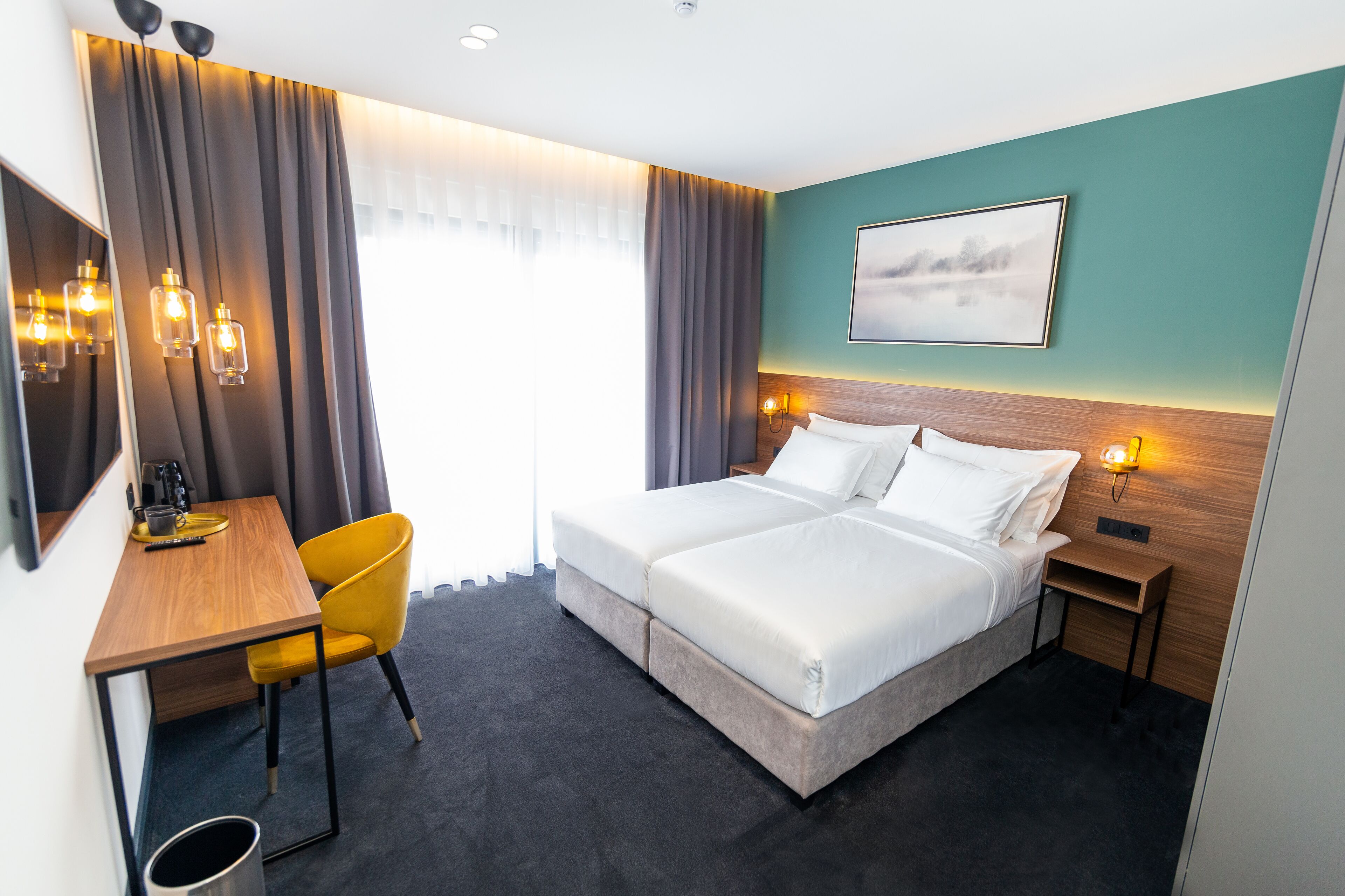 A hotel room with a double bed, a desk with a chair, a small table, and a nightstand with a lamp.