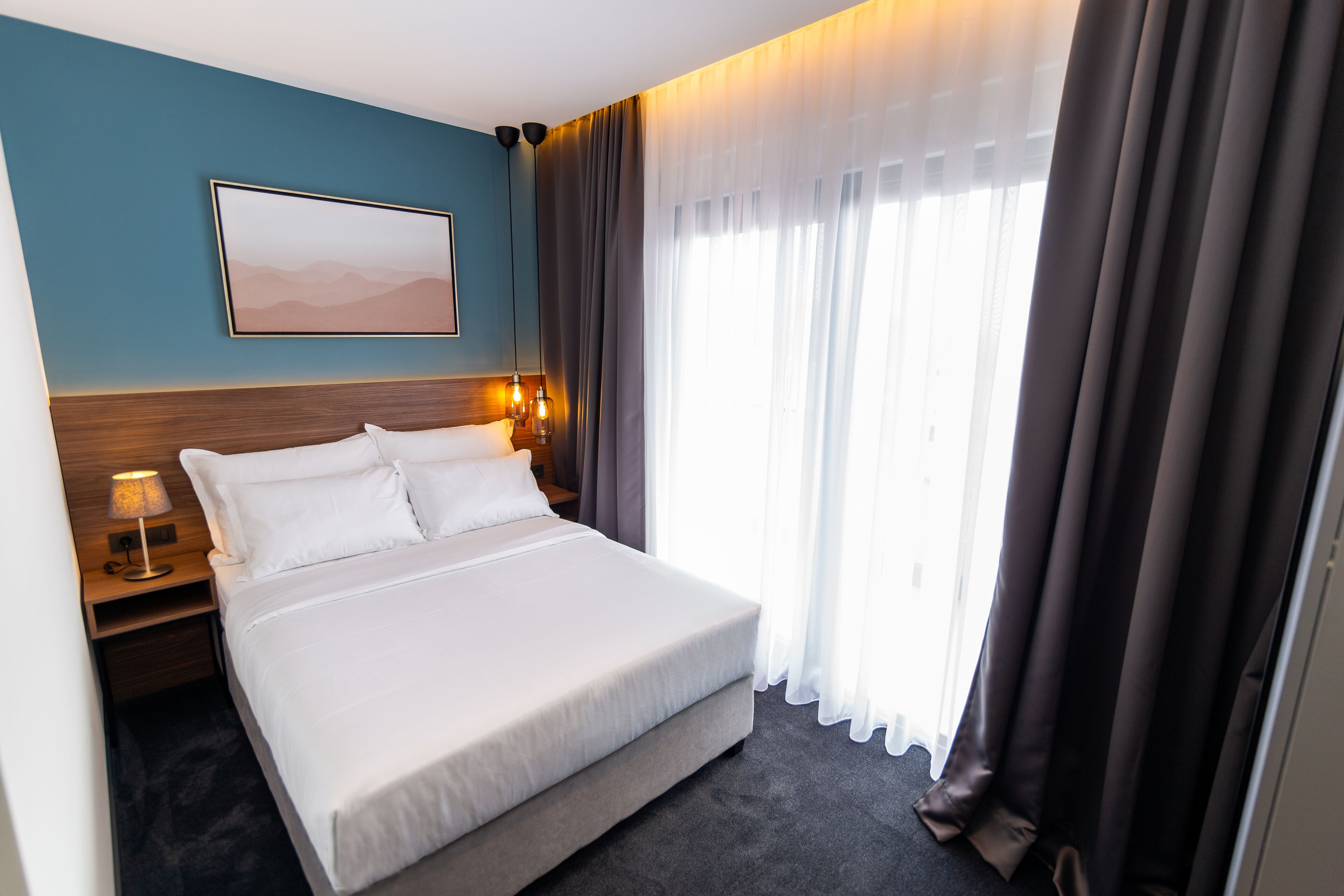 A hotel room with a large bed, a nightstand with a lamp, a wall-mounted picture, and a window with curtains.