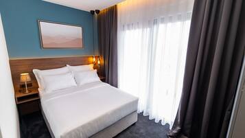 A hotel room with a large bed, a nightstand with a lamp, a wall-mounted picture, and a window with curtains.