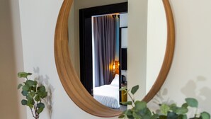 Una habitación con un espejo grande y redondo sobre una consola blanca, una cama con cortinas oscuras y un florero con plantas verdes.
