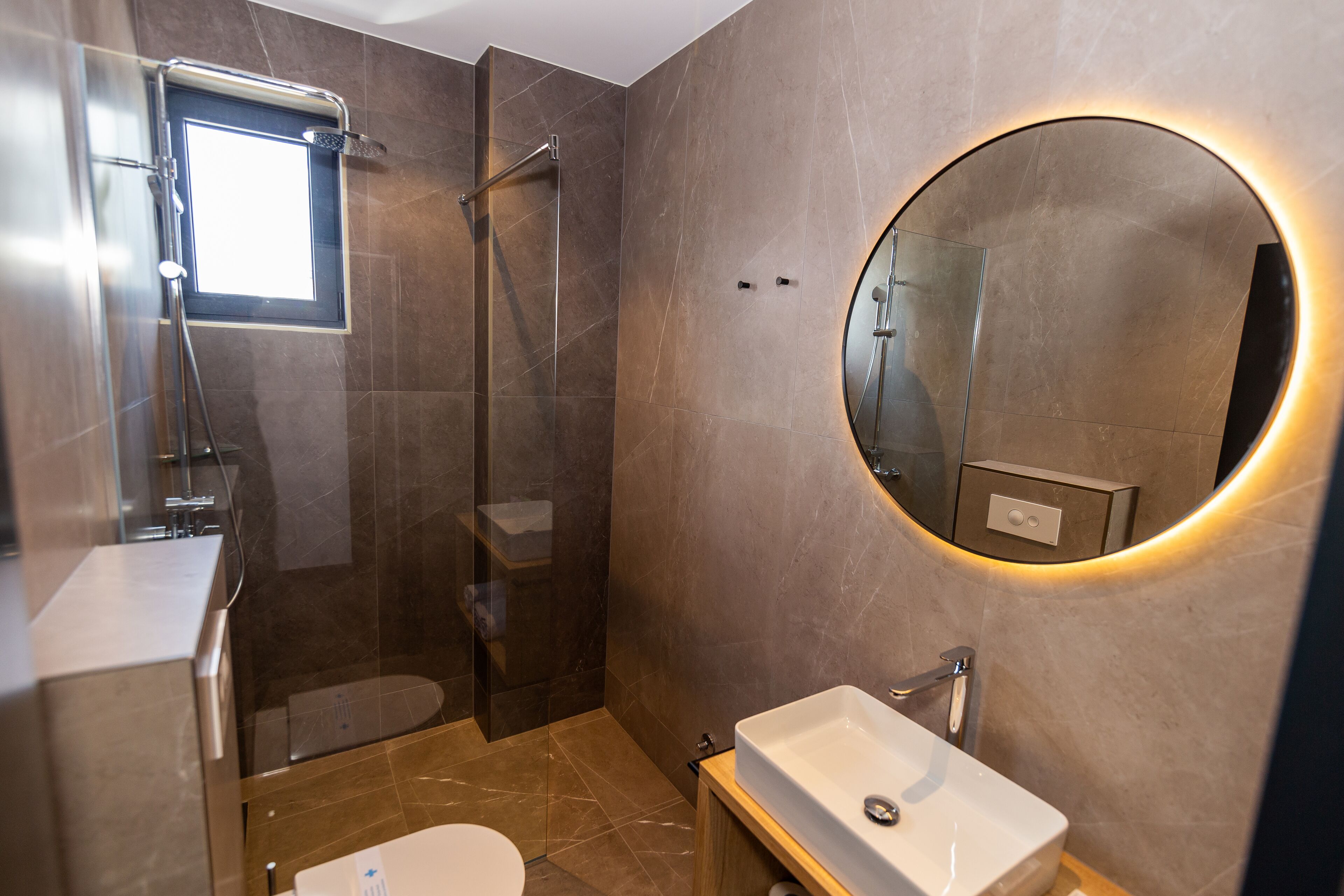 A modern bathroom with a glass-enclosed shower, a round mirror with a light halo, and a white sink.