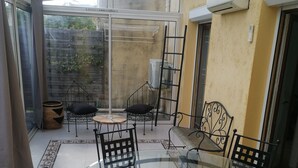 Une terrasse fermée par des parois de verre, avec une table ronde en verre et des chaises en métal noir, entourée d’un sol carrelé et d’un tapis tissé.