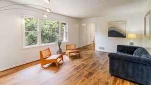 A spacious living room with hardwood flooring, a dark blue sofa, two wooden chairs, a large window with a view of trees, and a painting on the wall.