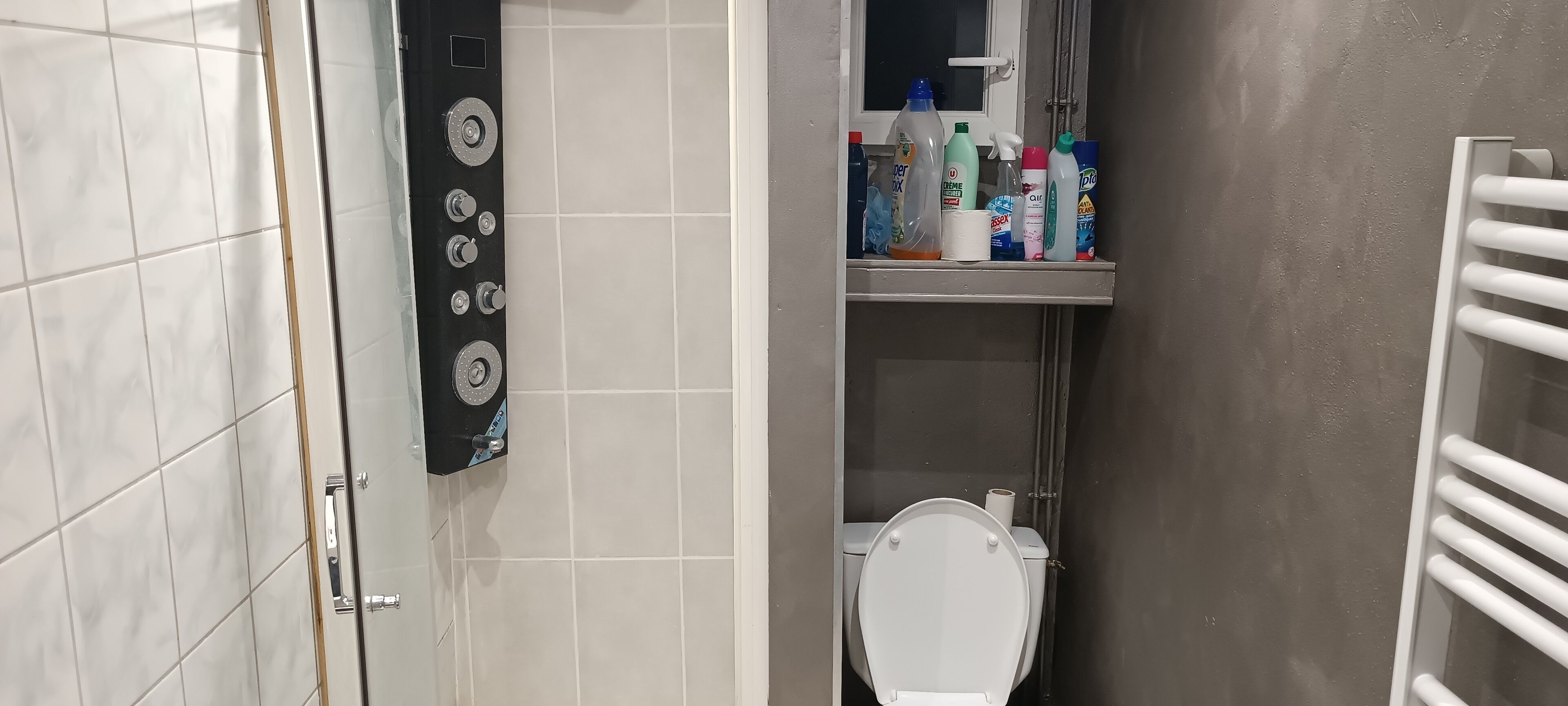 Un baño con inodoro, estante con productos de limpieza y panel de control.