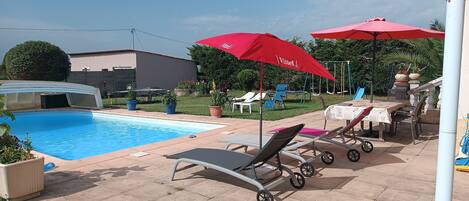 Uma área de piscina com espreguiçadeiras e guarda-sóis vermelhos, um espaço de estar coberto e um baloiço.