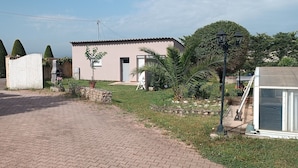 Une allée pavée mène à une maison avec un toit en tuiles, un mur en pierre et un jardin où poussent différentes plantes ainsi qu'un petit arbre.