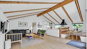 Geräumiges Loft mit hoher Decke, freiliegenden Holzbalken, einer gemütlichen Sitzecke mit Sofa und Sesseln, einer Küche mit Backsteinwand und Blick ins Grüne.