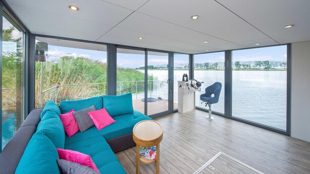 Une pièce moderne dotée d’une grande fenêtre donnant sur un plan d’eau, d’un canapé turquoise agrémenté de coussins, d’une table en bois et d’un panneau de commande.