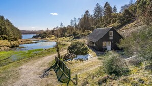 Een rustiek vakantiehuisje met een schuin dak, omgeven door een natuurlijke omgeving met een vijver en dichte bomen.