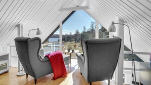 Een zolderkamer met twee grijze fauteuils, een rode plaid en uitzicht op een meer en bos.