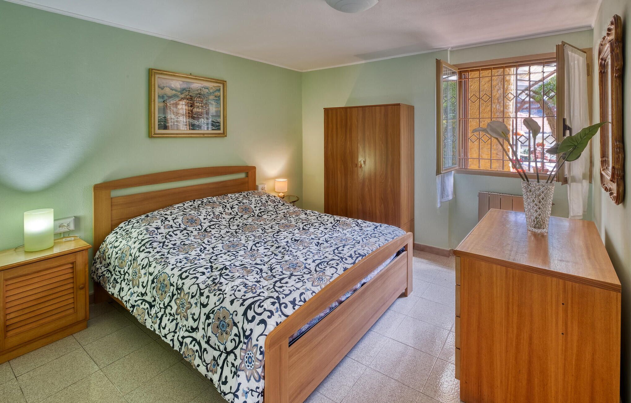 A bedroom with a bed, a nightstand, a wooden wardrobe, a window with a view, and a framed picture on the wall.