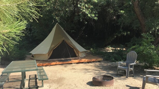 A tent with a wooden platform, a fire pit, and a table with chairs in a sandy outdoor area surrounded by trees. - Sequoia Dawn  (Three Rivers)