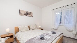 Quarto com cama, duas mesas de cabeceira, luminária e janela com cortinas leves.