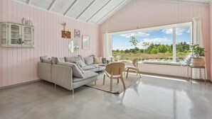 Un salon spacieux doté d’une grande fenêtre, d’un canapé gris, de chaises en bois et d’un meuble avec des portes en verre.