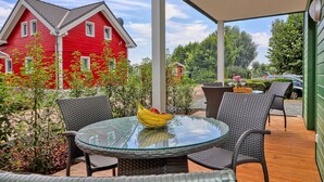 Ein Tisch mit Glasplatte, Korbstühle, ein rotes Haus im Hintergrund und eine überdachte Terrasse.