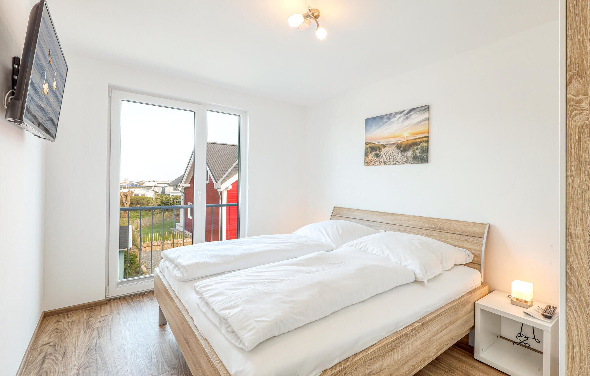 Een slaapkamer met een bed, nachtkastje, televisie, schilderij en balkon met uitzicht op gebouwen.