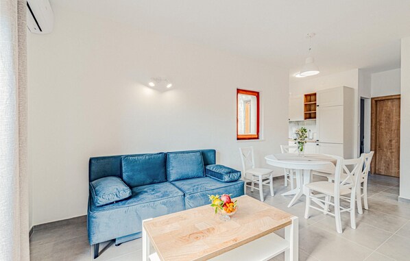 Una sala moderna con un sofá azul, una mesa de comedor blanca y sillas. Hay una pequeña área de cocina con armarios y una ventana.