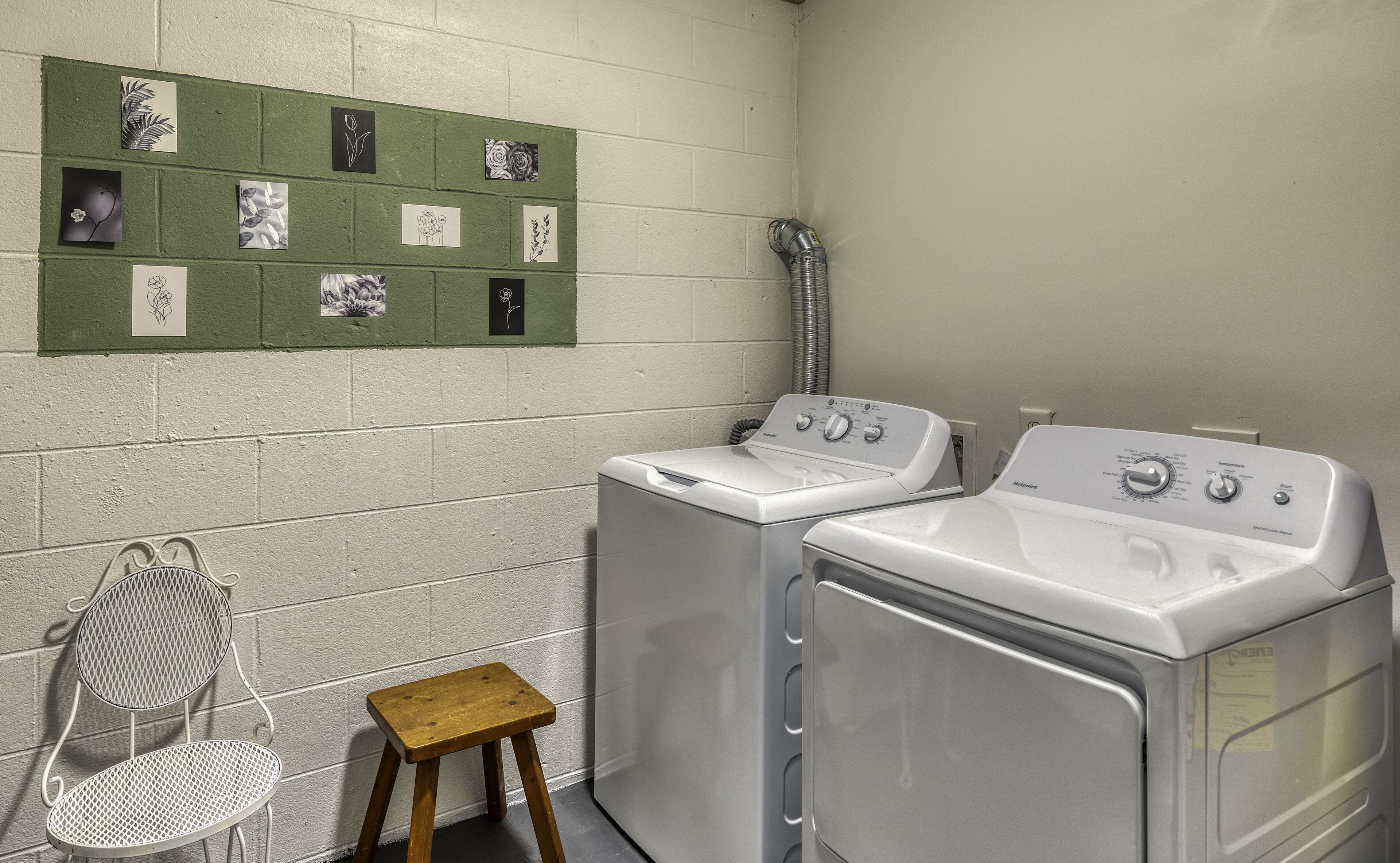 A laundry room with two white front-loading washing machines, a small wooden stool, a metal chair, and a wall-mounted drying rack with floral designs.