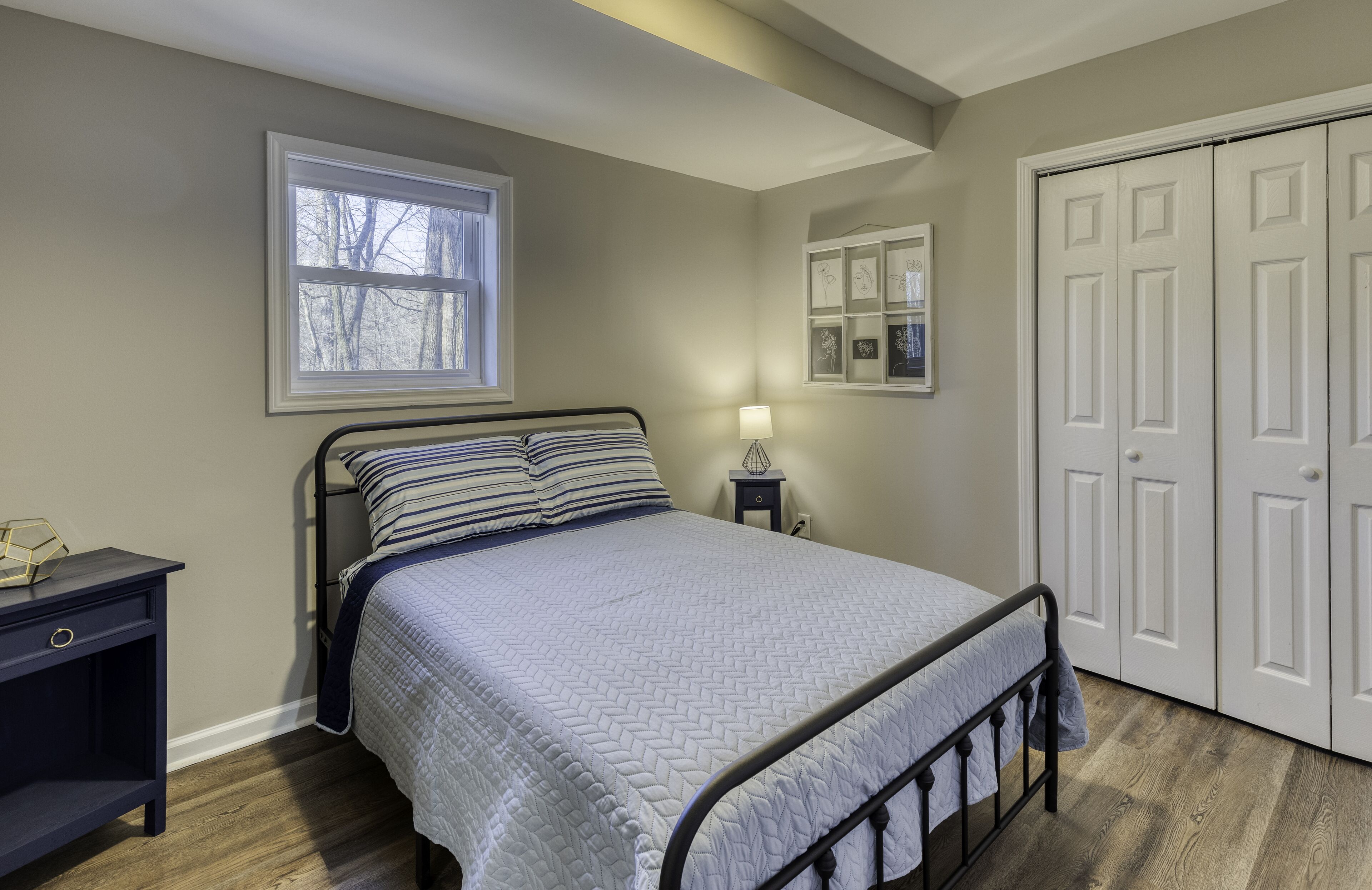 A bedroom with a bed, a nightstand, a window, a lamp, and a set of white doors.