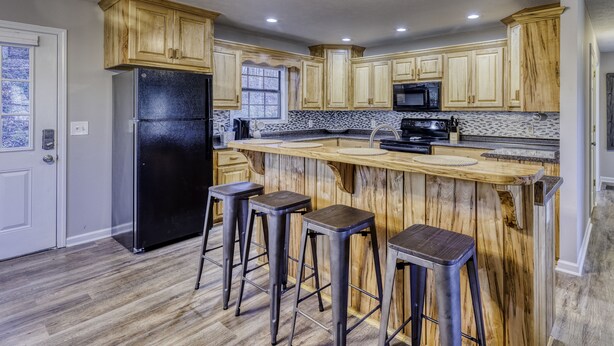 A kitchen with wooden cabinets, a black refrigerator, a microwave, and a bar area with stools. - Lake, Fun, BBQ & Family. Charming & Cozy HOME! (morganton)