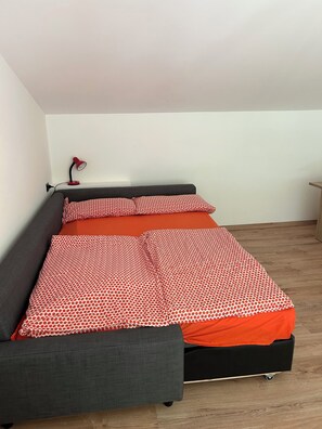Ein Einzelbett mit grauem Gestell, rot-weißer Bettwäsche und einer roten Schreibtischlampe.
