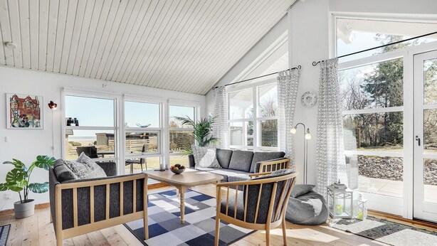A bright, open-plan living area with a wooden floor, a sofa set, a dining table, and large windows offering views of the outdoors.