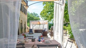 Un espace extérieur aménagé pour les repas, avec une table dressée pour deux personnes, entourée de chaises, un coin salon abrité et un bâtiment en arrière-plan.