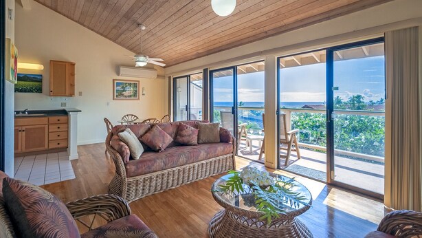 A spacious living area with a large wicker sofa, a glass coffee table with flowers, and a view of the ocean through large sliding glass doors. - 180' view 🌊& Mountain View! 1000 feet to 🏝watch 🌅 (Koloa)