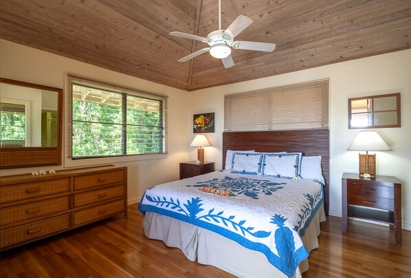 A bedroom with a wooden ceiling, a bed with a floral bedspread, a nightstand with a lamp, a dresser, and a window with blinds.