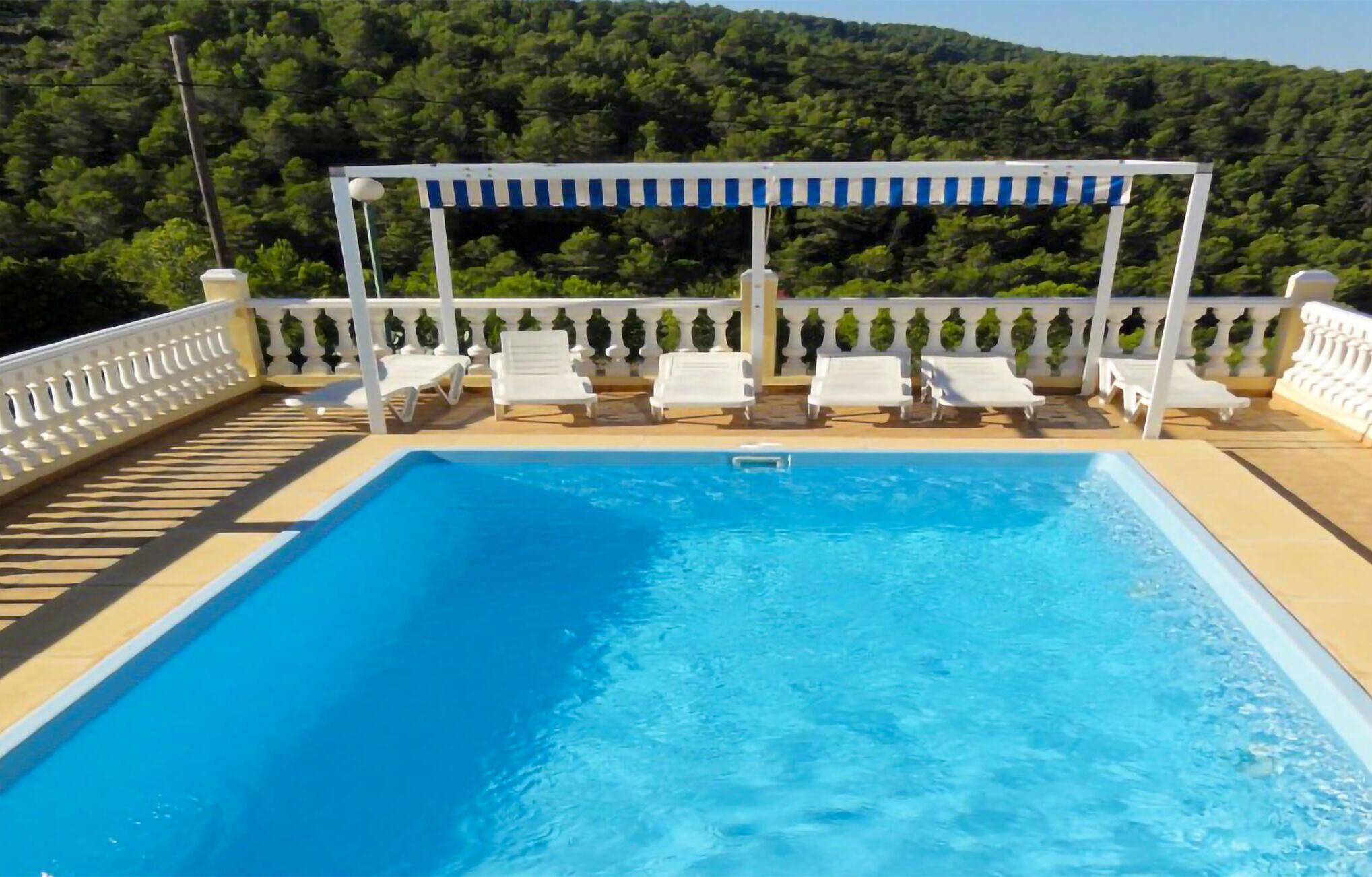 Une piscine rectangulaire entourée de chaises longues blanches, une pergola surmontée d’une marquise à rayures et une balustrade blanche.