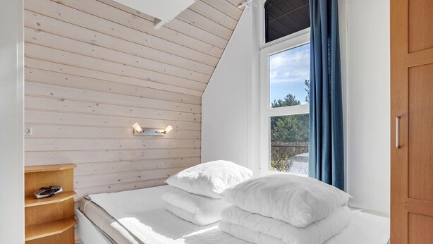 A bedroom with a sloped ceiling, a bed with white bedding, a wooden shelf, and a window with curtains. - Gorgeous home in Hadsund with WiFi (Hadsund)