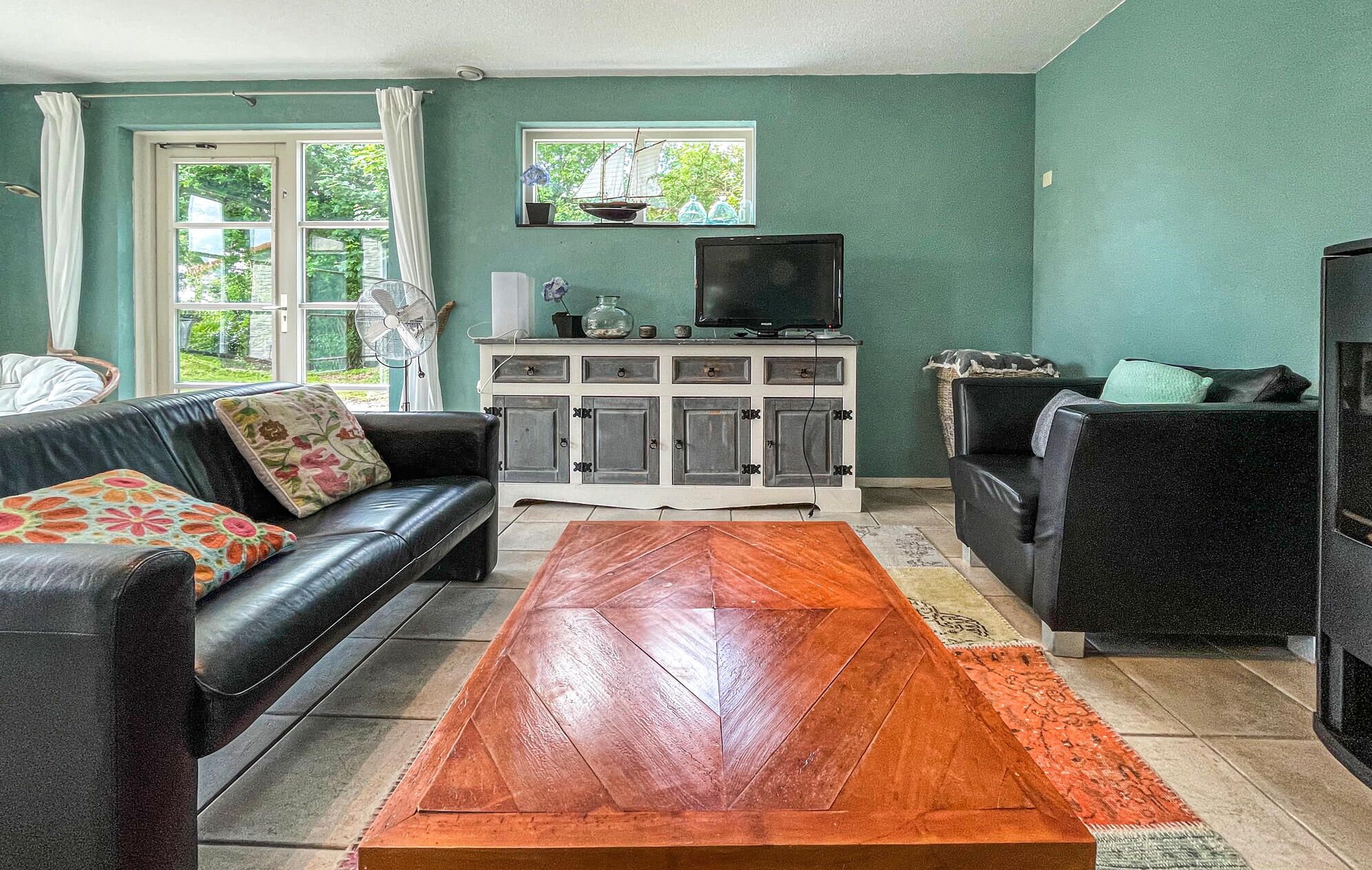 A living room with a black leather sofa, a wooden coffee table, a TV on a white cabinet, and a window with curtains.