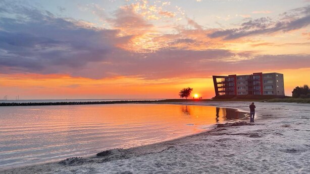 A beach at sunset with a person standing on the sand, a building in the background, and a calm sea reflecting the sky. - Cozy home in Makkum with kitchen (Makkum)